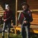 Several musicians play the accordion, harmonica, washboard and other instruments on a log cabin porch
